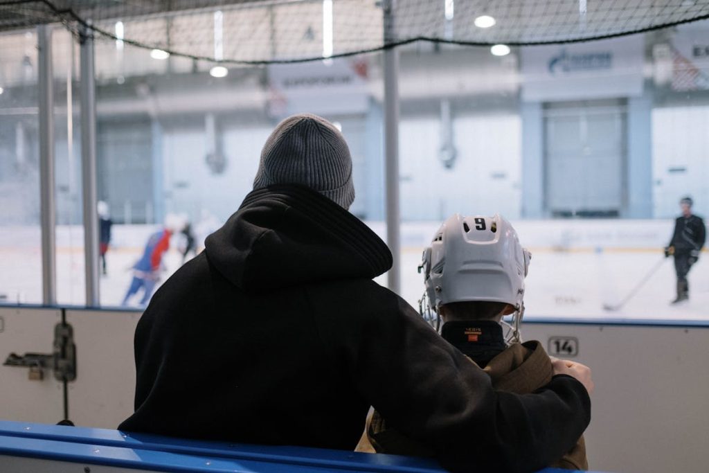 A parent and child in ice hockey gear watching a game at an indoor rink.