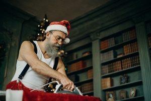 Bearded man dressed as Santa Claus ironing clothes in a festive library setting with Christmas decor.