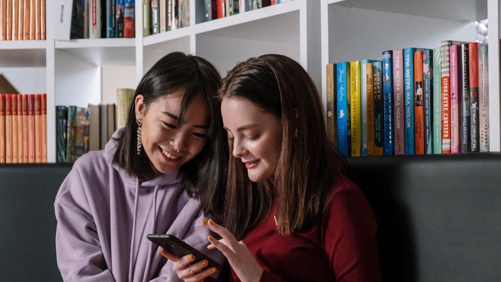 Two teenage girls sitting in a library sharing a mobile phone, smiling and enjoying their time.