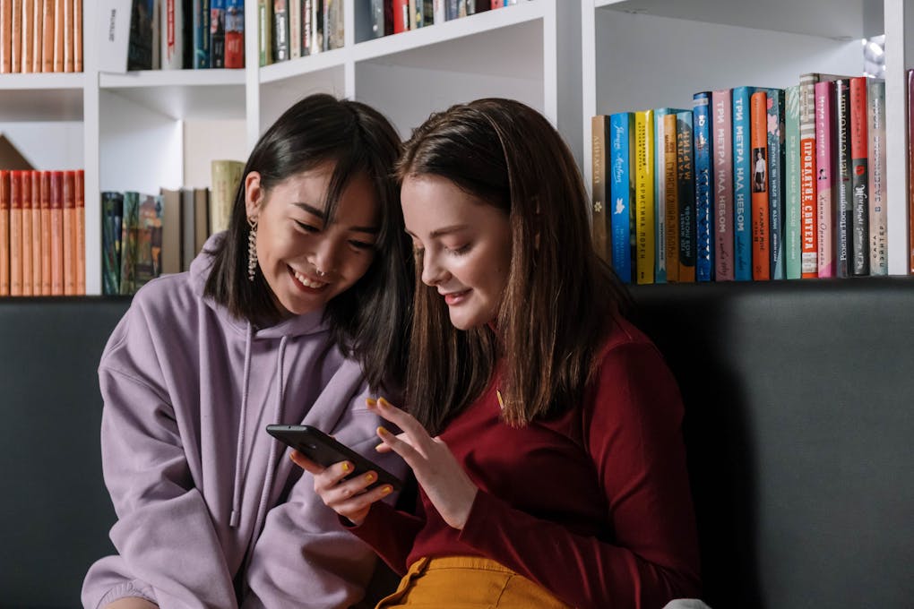 Two teenage girls sitting in a library sharing a mobile phone, smiling and enjoying their time.